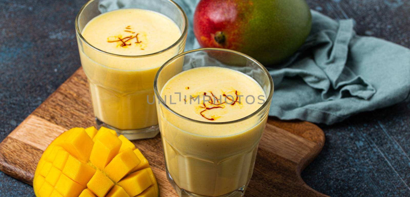 Healthy Indian Ayurveda drink mango lassi in two glasses on rustic concrete table with fresh ripe cut mango, yellow blended beverage made of mango fruit, yoghurt or milk curd and spices