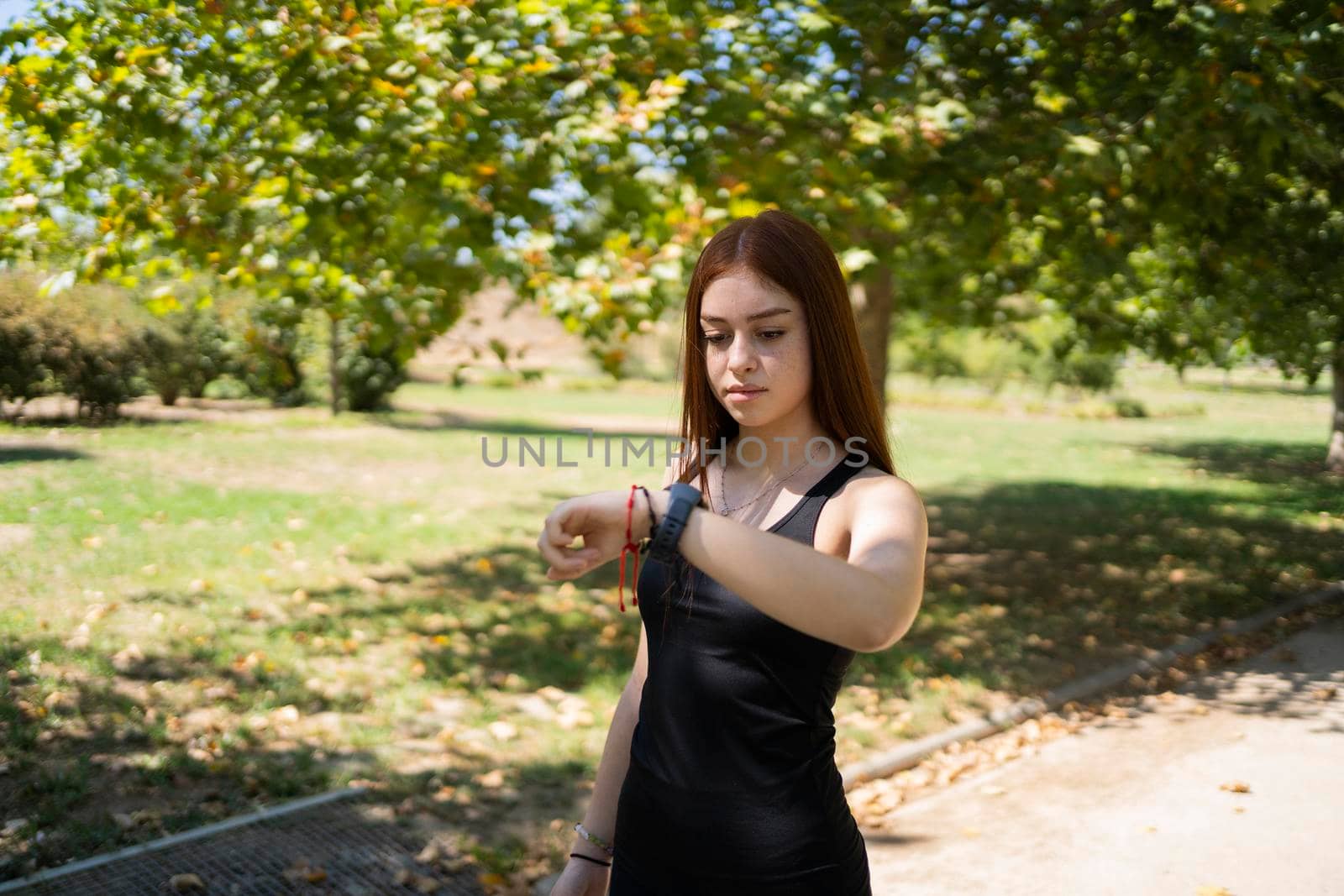 Young sportswoman checking pulse on smart watch while standing on sunlit alley during break in running session in park