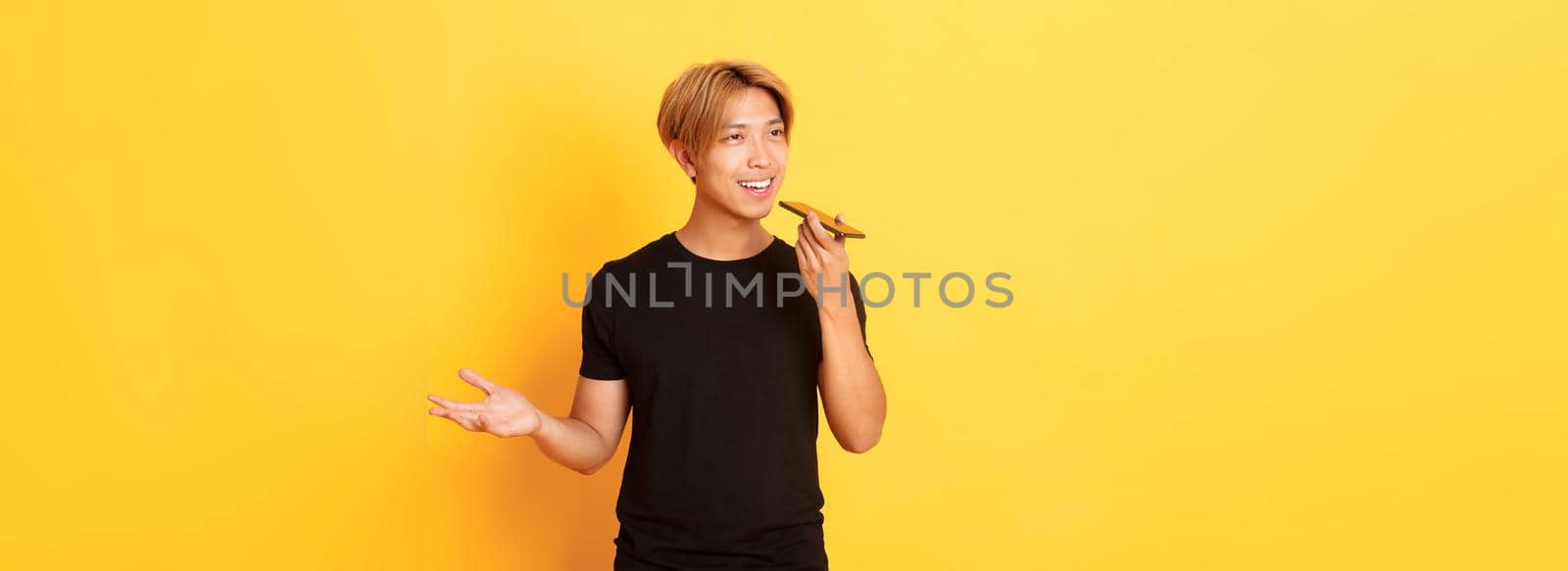 Portrait of stylish handsome korean guy with blond hair record voice message on mobile phone, holding smartphone near mouth and speaking, standing yellow background.