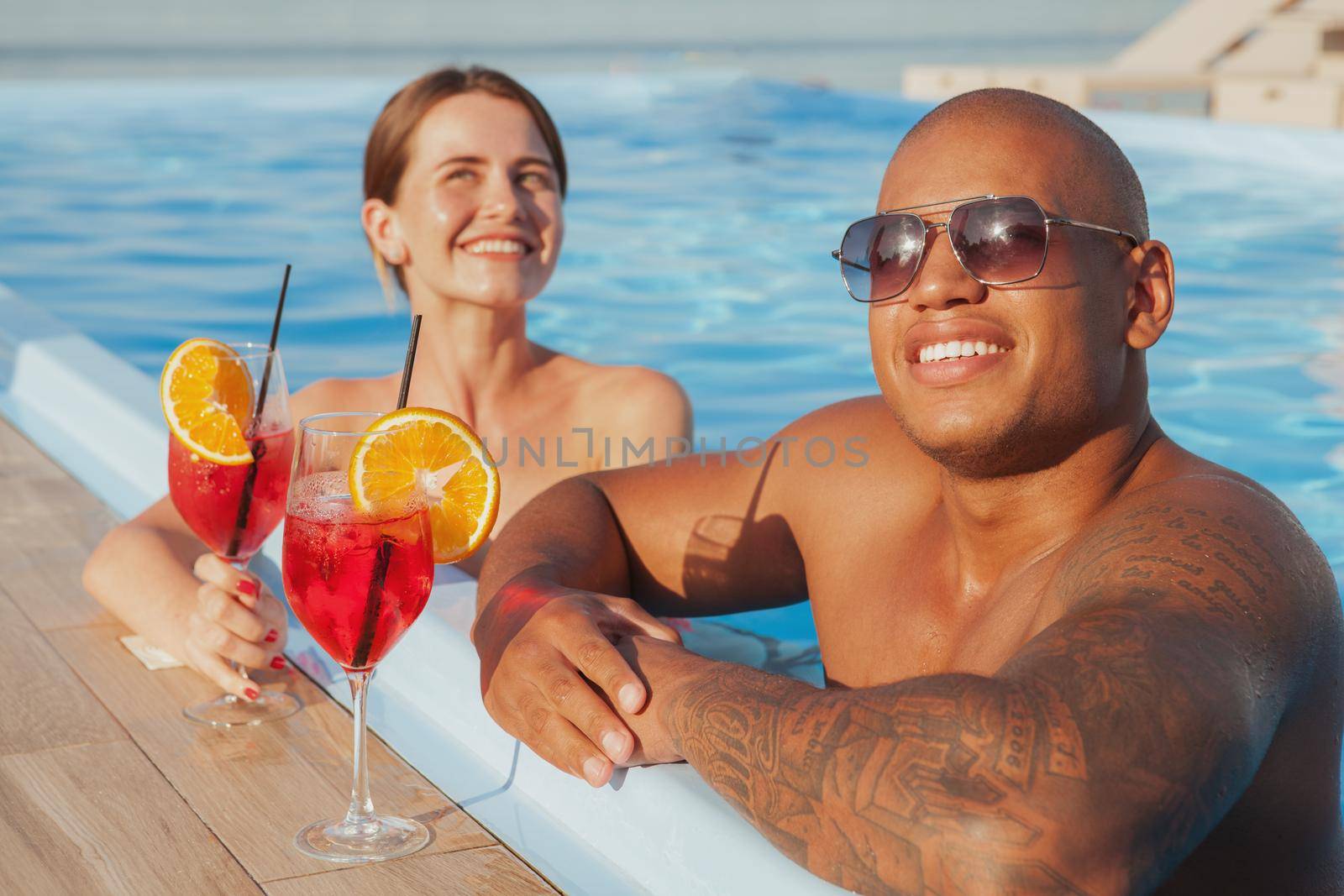 Happy handsome African man and his girlfriend relaxing in the swimming pool, enjoying their summer vacation. Seaside, resort, hotel concept