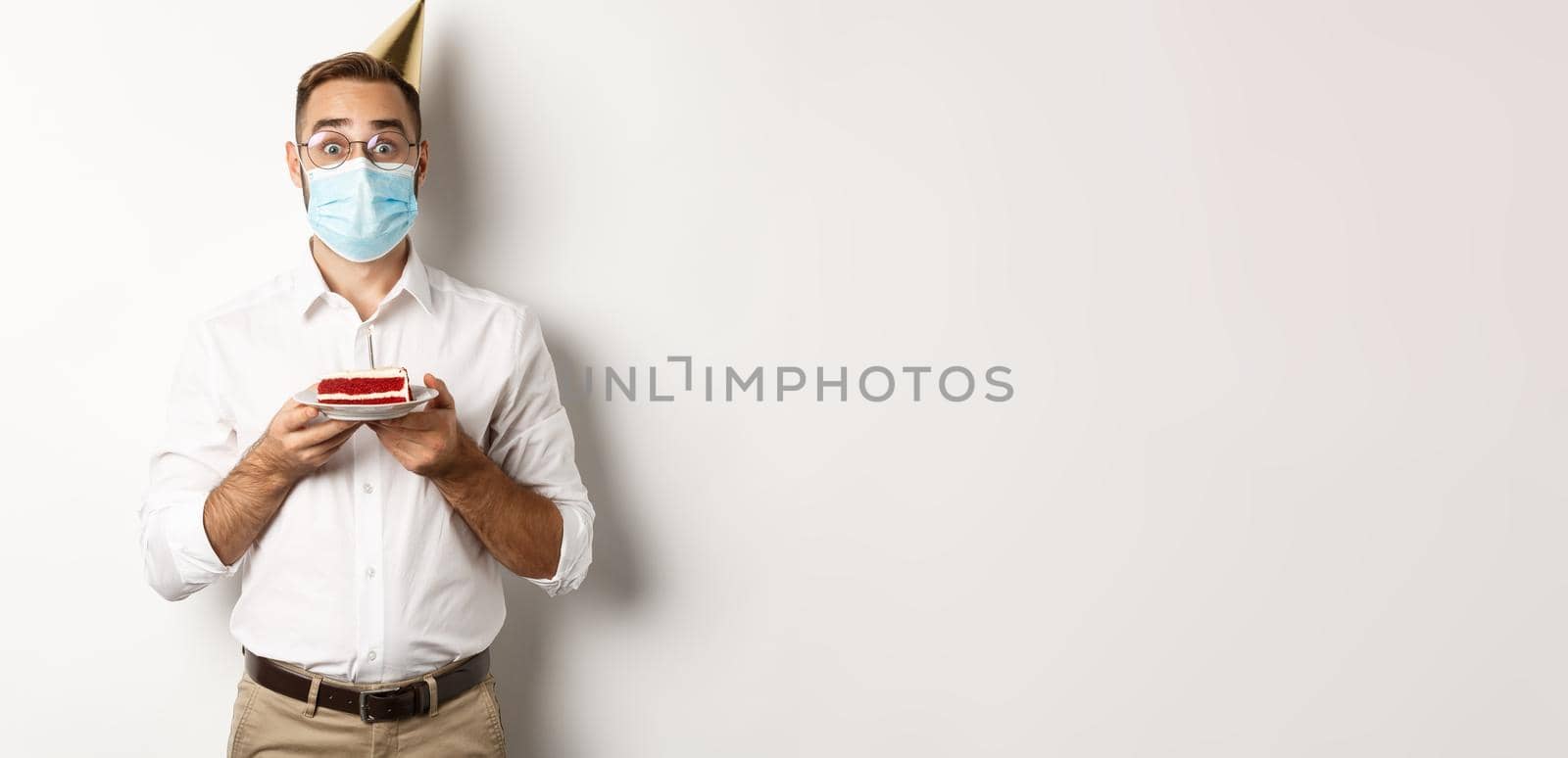 Covid-19, social distancing and celebration. Surprised birthday guy holding bday cake, wearing face mask from coronavirus, white background.