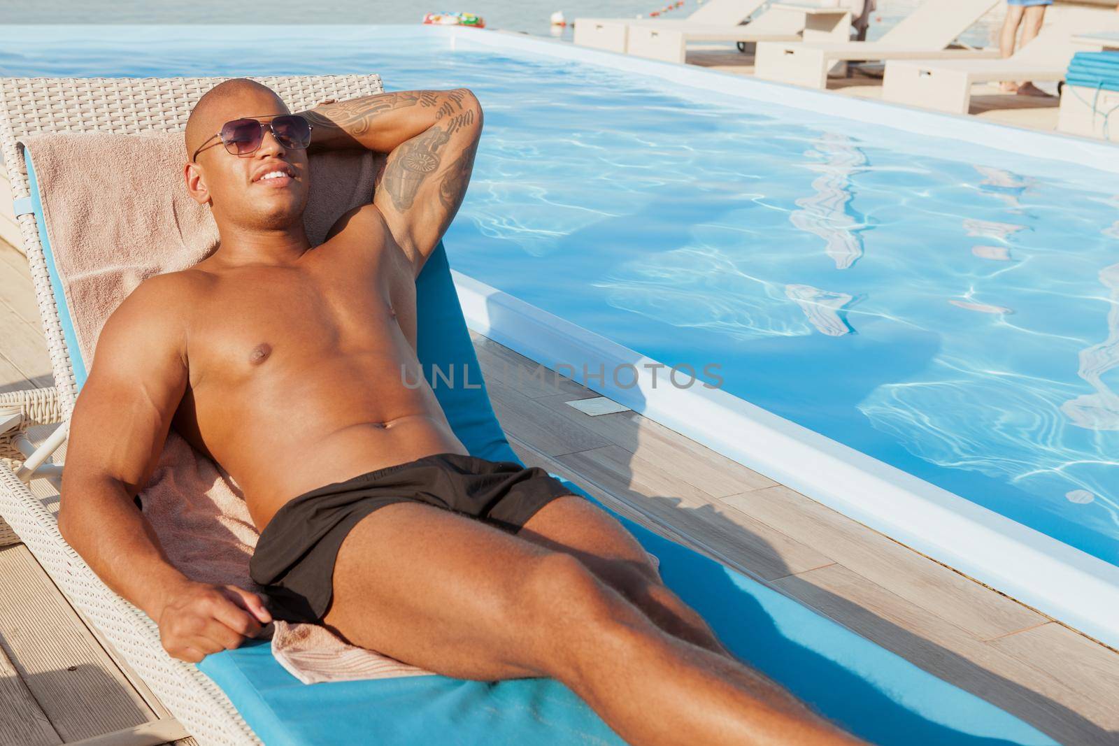 Relaxed African man with athletic body sunbathing at the poolside, copy space. Handsome fit young man resting near the swimming pool, enjoying his vacation