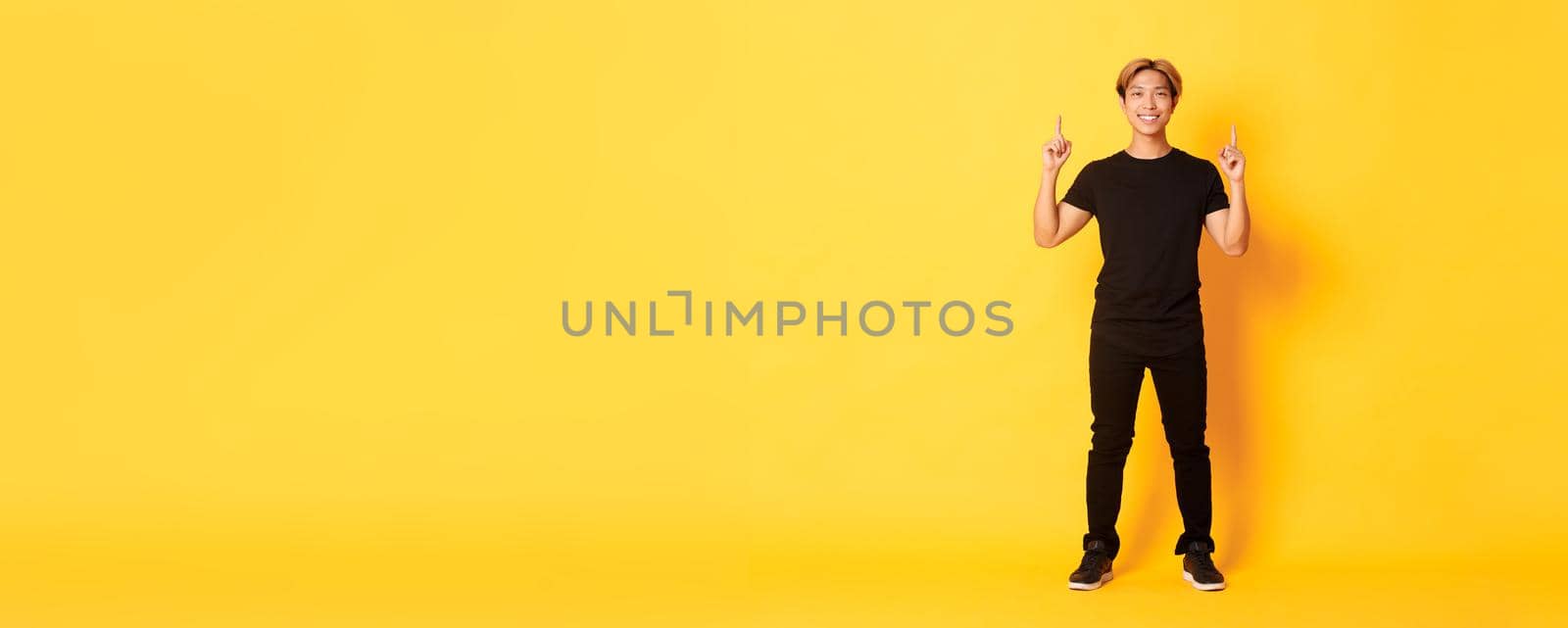 Full-length of confident handsome asian man with blond hair, smiling and pointing fingers up, showing logo, yellow background by Benzoix