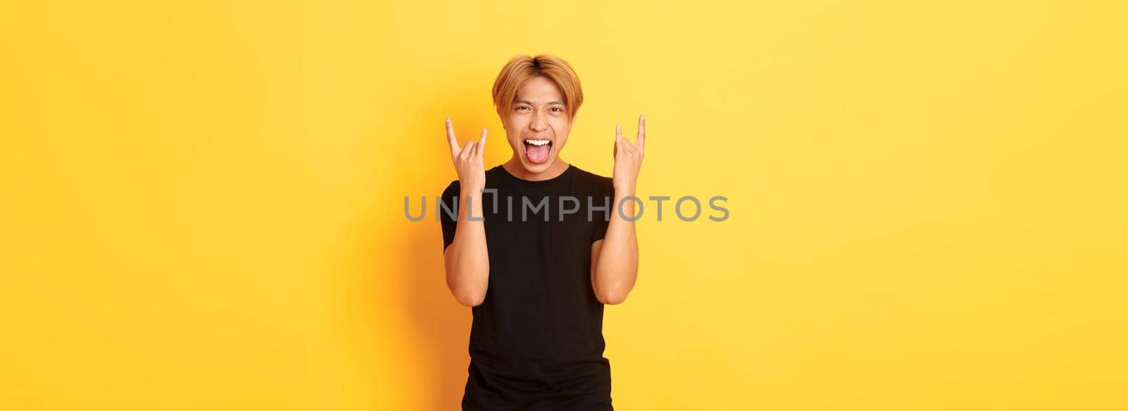 Portrait of joyful handsome asian blond guy having fun, showing rock-n-roll gesture and stick tongue carefree, standing yellow background by Benzoix