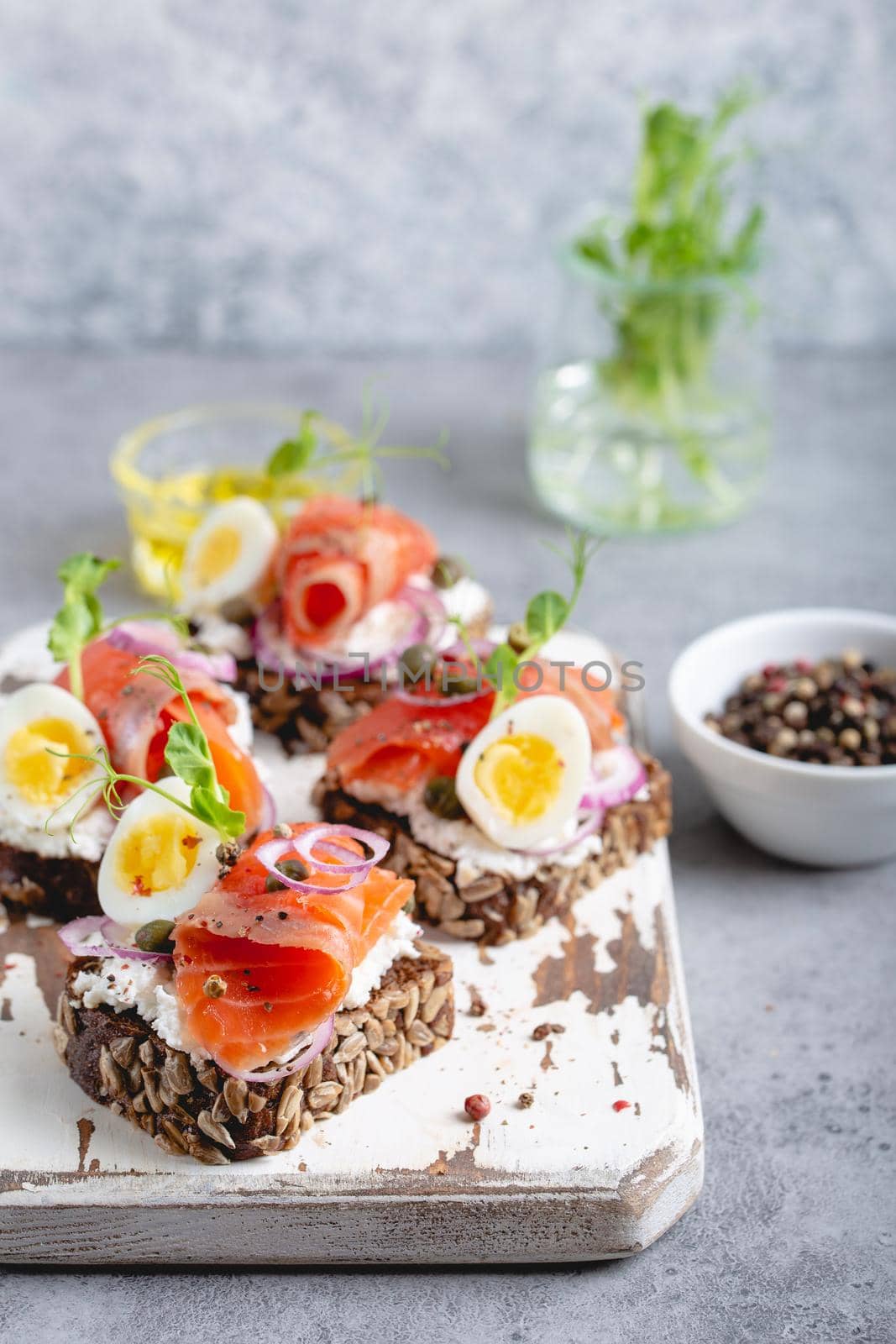 Close-up of sandwich with dark rye bread, cream cheese, salmon, onion, capers, boiled egg on white wooden cutting board, concrete background. Traditional Scandinavian open sandwich Smorrebrod.