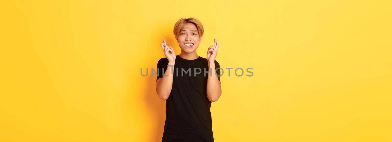 Portrait of nervous asian guy cross fingers good luck, making wish, praying over yellow background by Benzoix