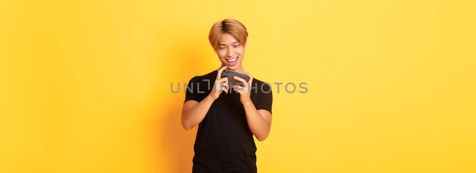 Portrait of stylish handsome asian guy playing smartphone game, looking amused at mobile screen, standing yellow background by Benzoix