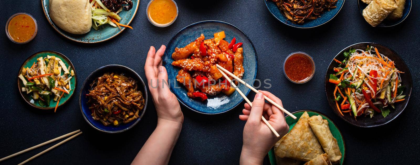 Set of Chinese dishes on table, female hands holding chopsticks: sweet and sour chicken, fried spring rolls, noodles, rice, steamed buns with bbq glazed pork, Asian style banquet or buffet, top view .