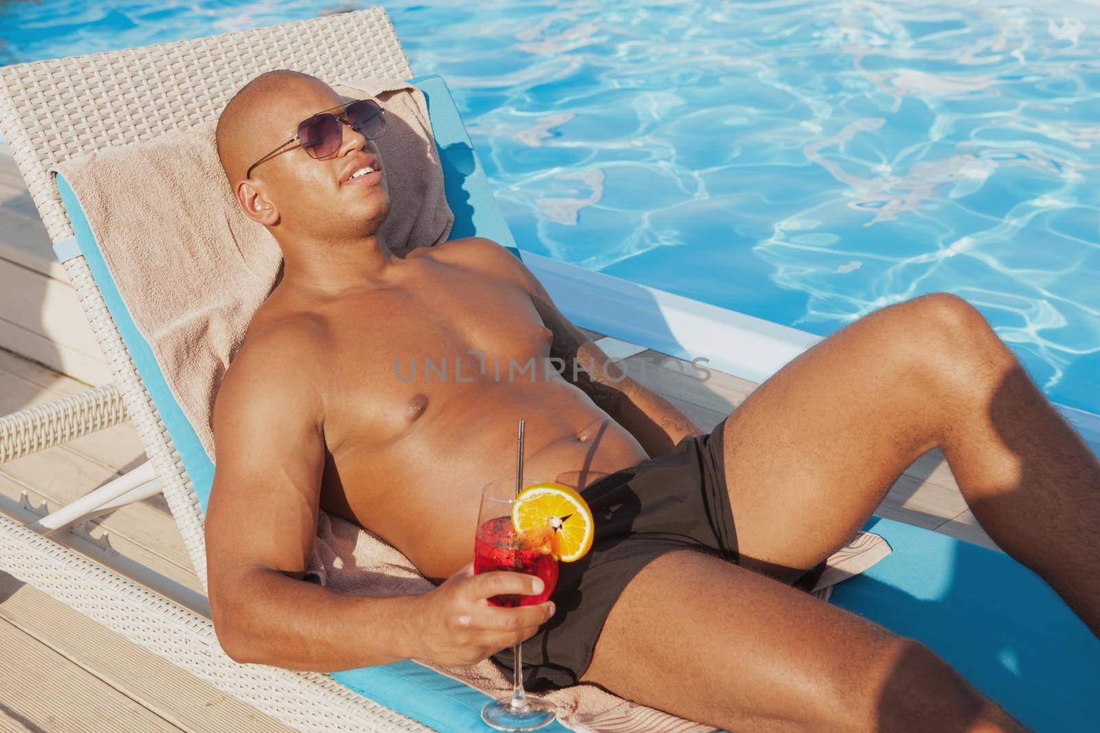 Attractive relaxed African man enjoying sunbathing, having a drink at the poolside. Handsome athletic man tanning near the swimming pool, copy space