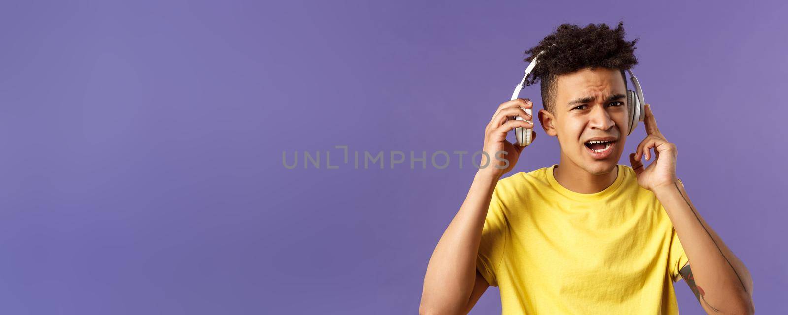 Cant hear you, repeat please. Portrait of young bothered guy interrupted of listening music, take-off headphones to answer person question, squinting look confused, purple background.