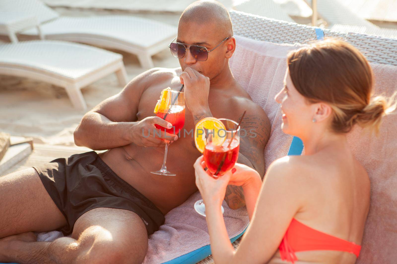 Happy young multiethnic couple relaxing at the beach together, having cocktails. Athletic African man sipping his drink, enjoying summer vacation at the seaside with girlfriend