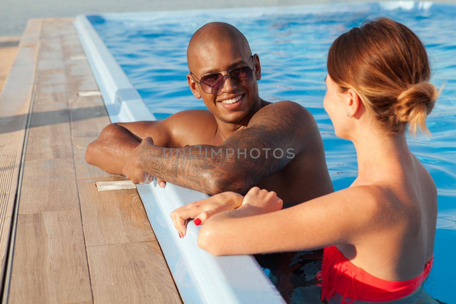 Attractive cheerful African man talking to his girlfriend at the swimming pool. Multiethnic couple enjoying summer vacation together, resting after swimming at resort poolside