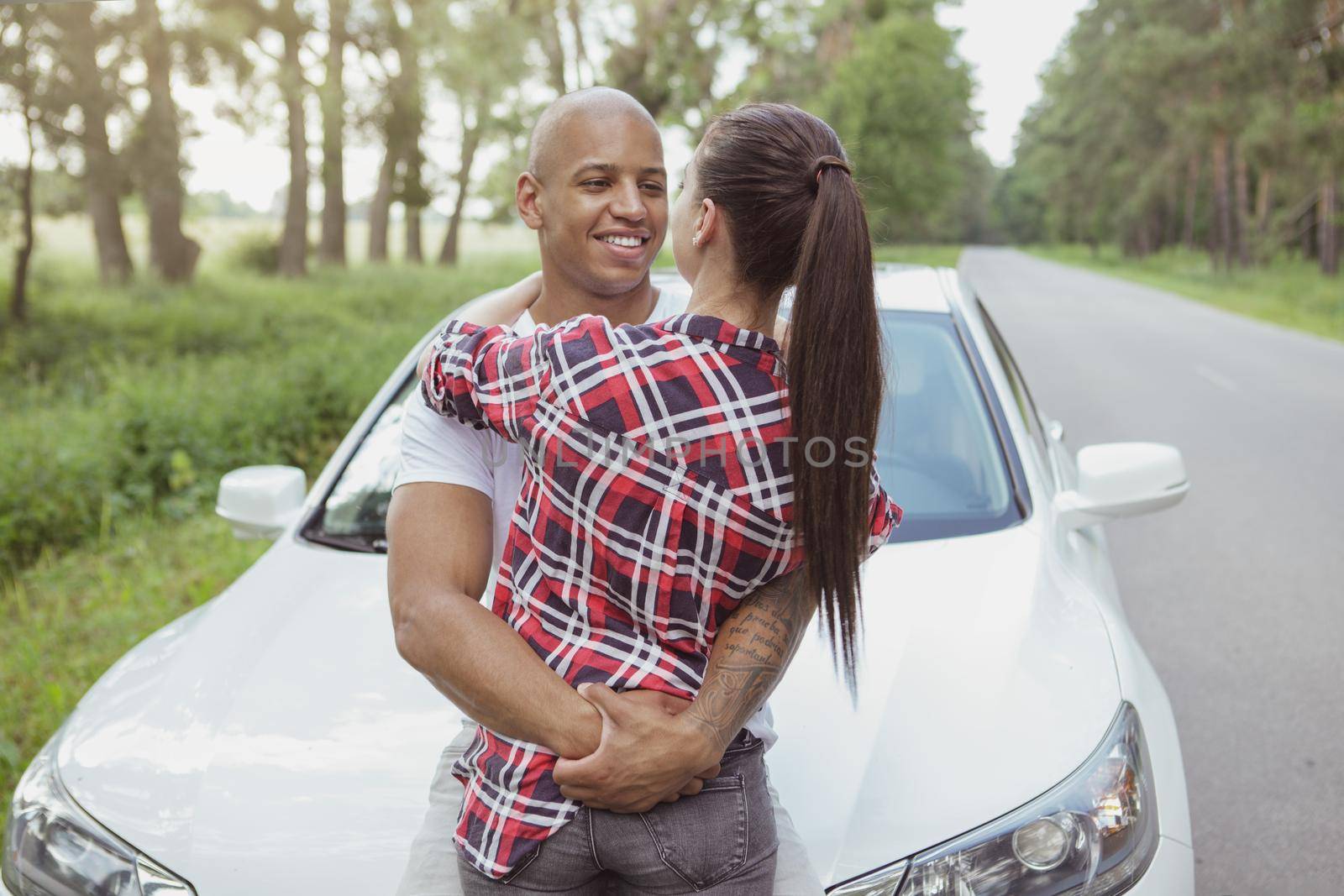 Handsome happy African man embracing his girlfriend near the car, while on a roadtrip together. Loving young couple enjoying travelling by car together