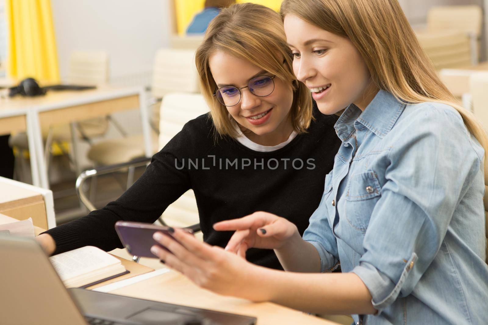 Beautiful young woman showing something on her smart phone to a female friend sitting together in front of the laptop at the office designers creative team working studying friendship mobility carrier