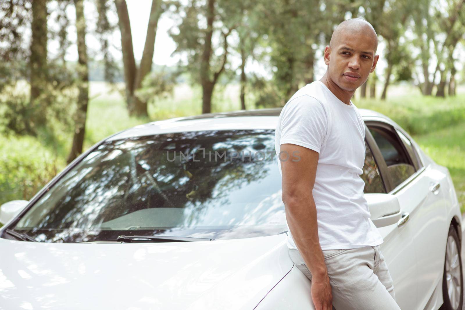 Attractive African man leaning on his car on a countryside road, looking away thoughtfully, copy space. Handsome male driver resting from drving during road trip