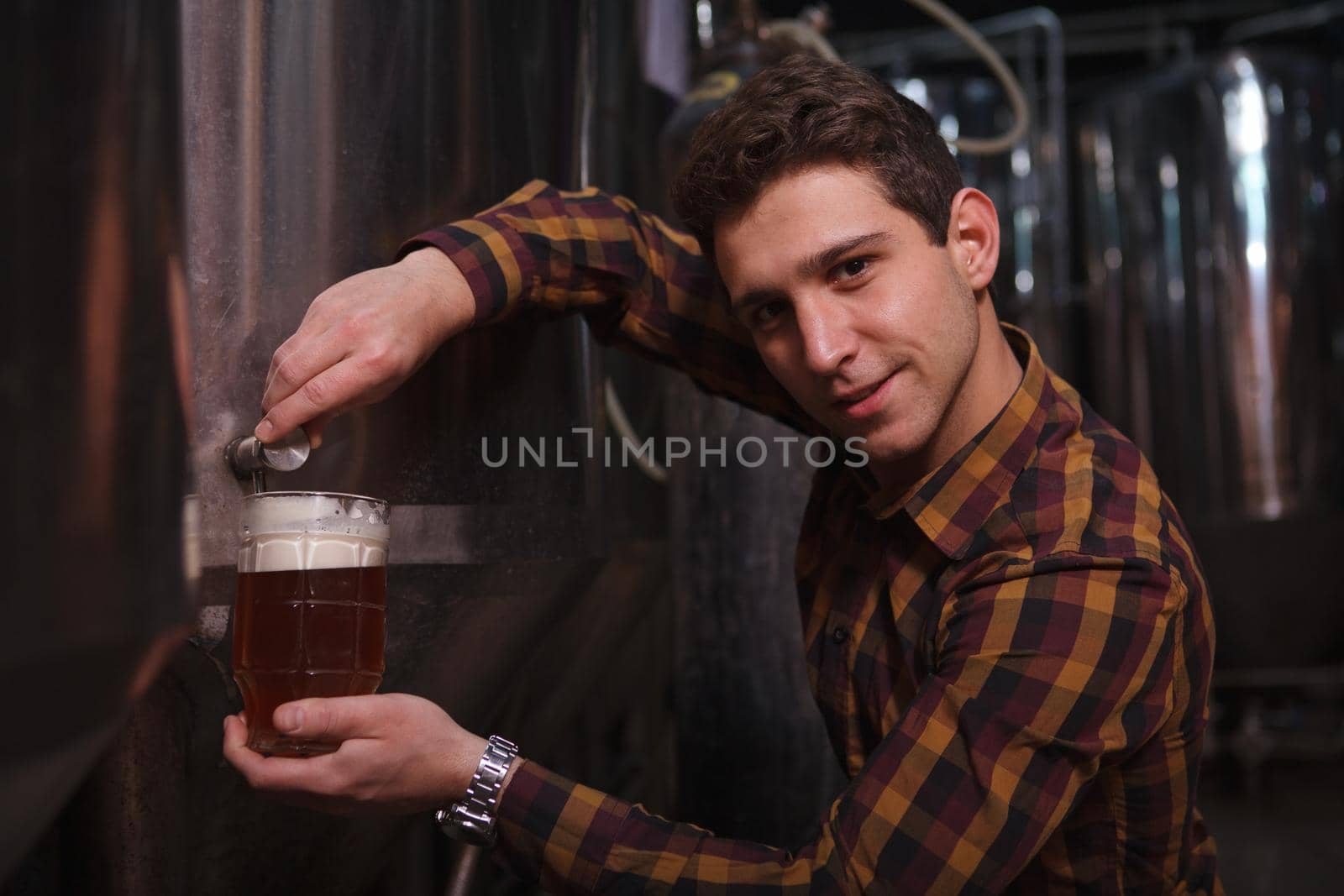 Handsome cheerful brewer looking to the camera while pouring freshly brewed beer from the tank into a mug at his microbrewery