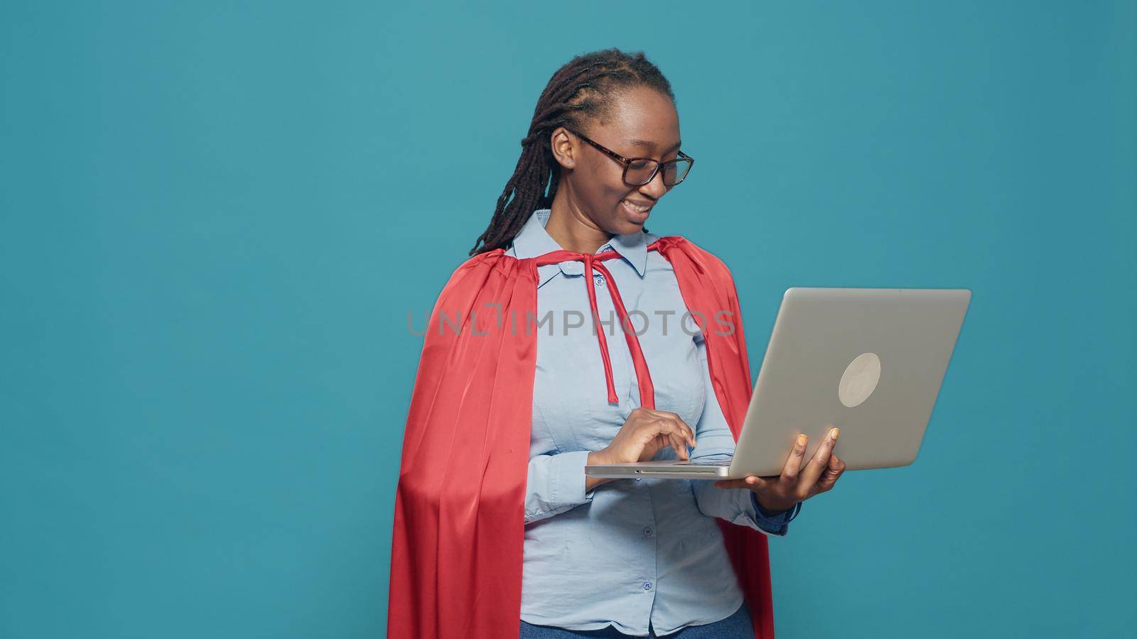 Woman dressed as superhero using laptop in studio, wearing red cape hero costume and browsing internet. Looking at computer with online network, having cloak and cartoon comic look.