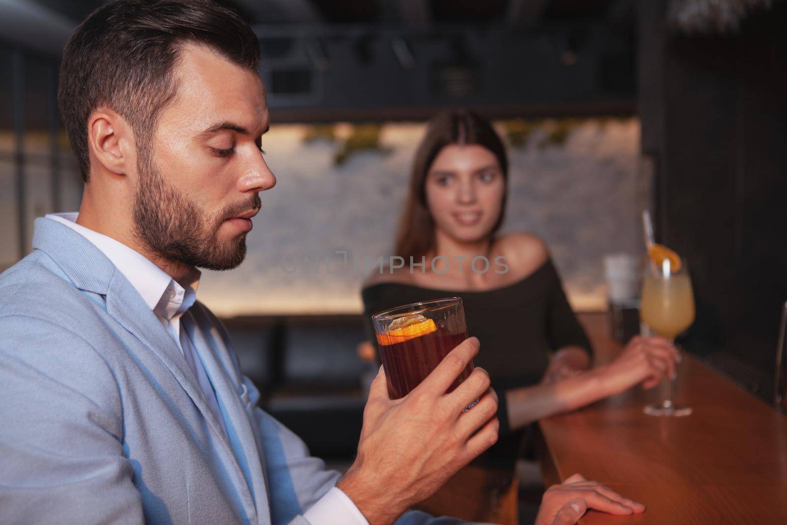 Handsome man drinking whiskey cocktail, woman looking at him seductively. Attractive bachelor enjoying a drink. Flirt, romance, seduction concept