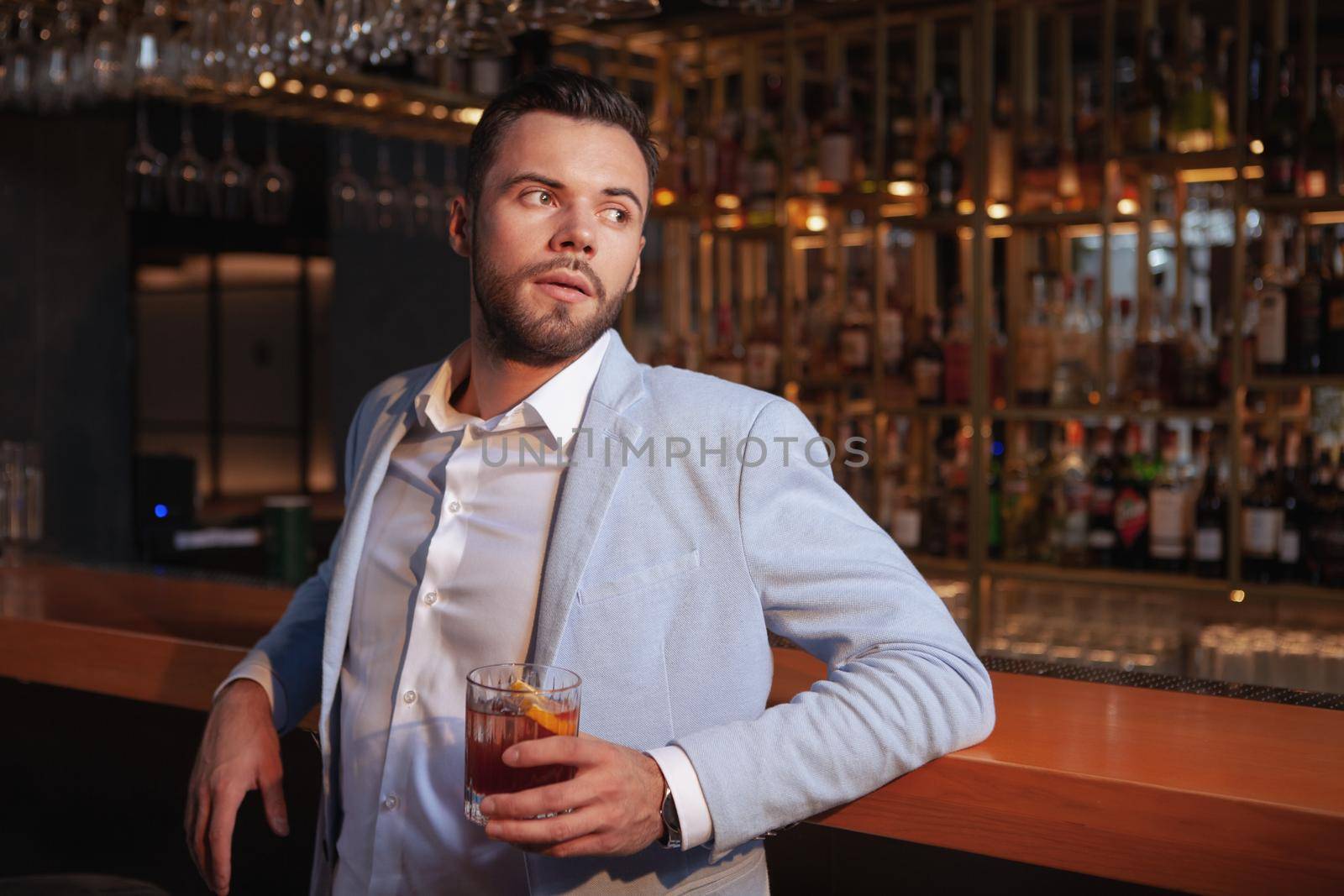 Handsome elegant young man relaxing at cocktail bar, leaning on bar counter, copy space