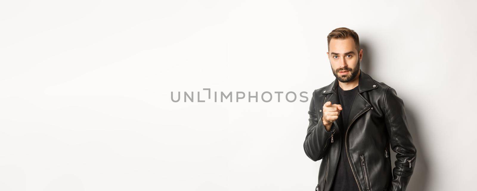 Handsome and cool bearded guy pointing finger at camera, wearing leather jacket, standing sassy against white background.
