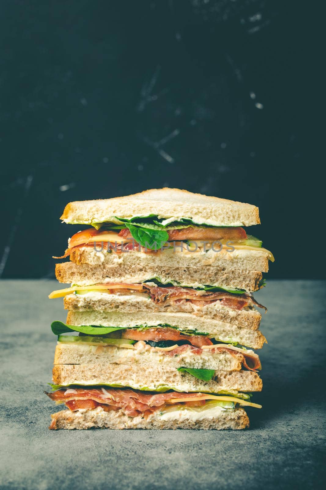 Close up of tall cut tasty sandwich with cheese, ham, prosciutto, fresh lettuce, tomatoes, cucumbers on gray rustic stone background. Healthy gourmet sandwich for breakfast or lunch, concept