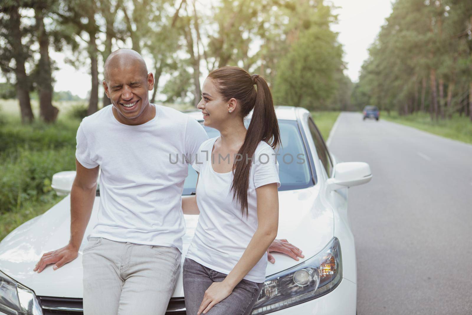 Lovely young couple laughing, enjoying their roadtrip on a car, copy space. Handsome African man laughing, embracing his girlfriend, leaning on a car on the side of the road