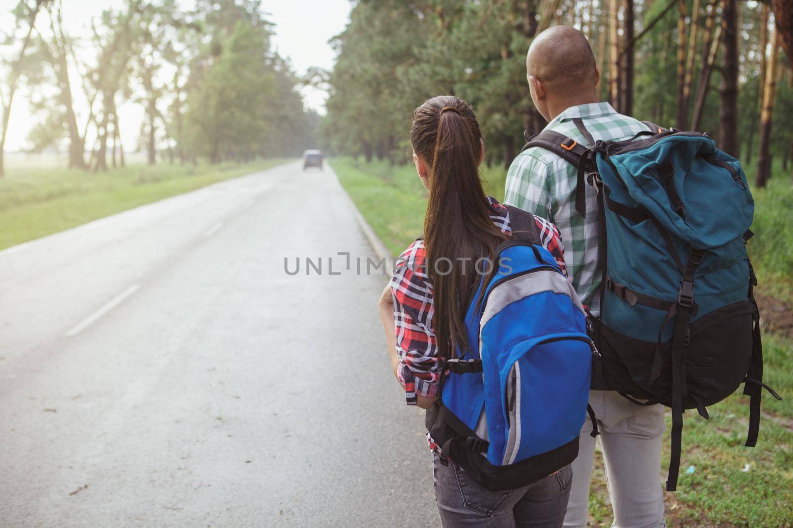 Rear view shot of a mixed couple with backpacks hitchhiking on countryside road in the forest, copy space