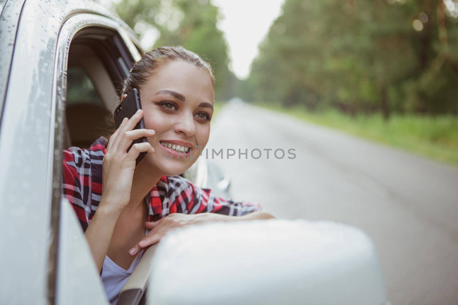 Beautiful happy young woman smiling, calling someone via her smart phone while on a roadtrip. Attractive woman enjoying travelling by car, using her mobile phone, copy space