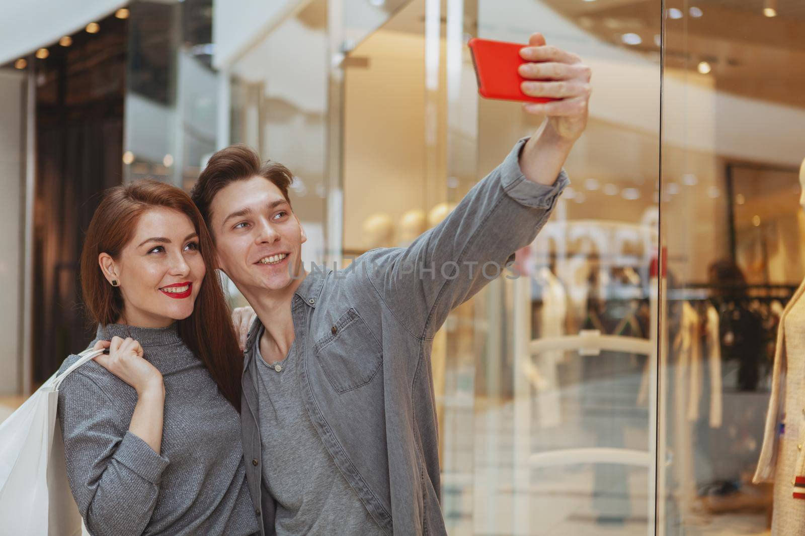 Handsome cheerful man taking selfies with his beautiful girlfriend, using smart phone at the shopping mall, copy space. Technology, social media, blogging lifestyle concept
