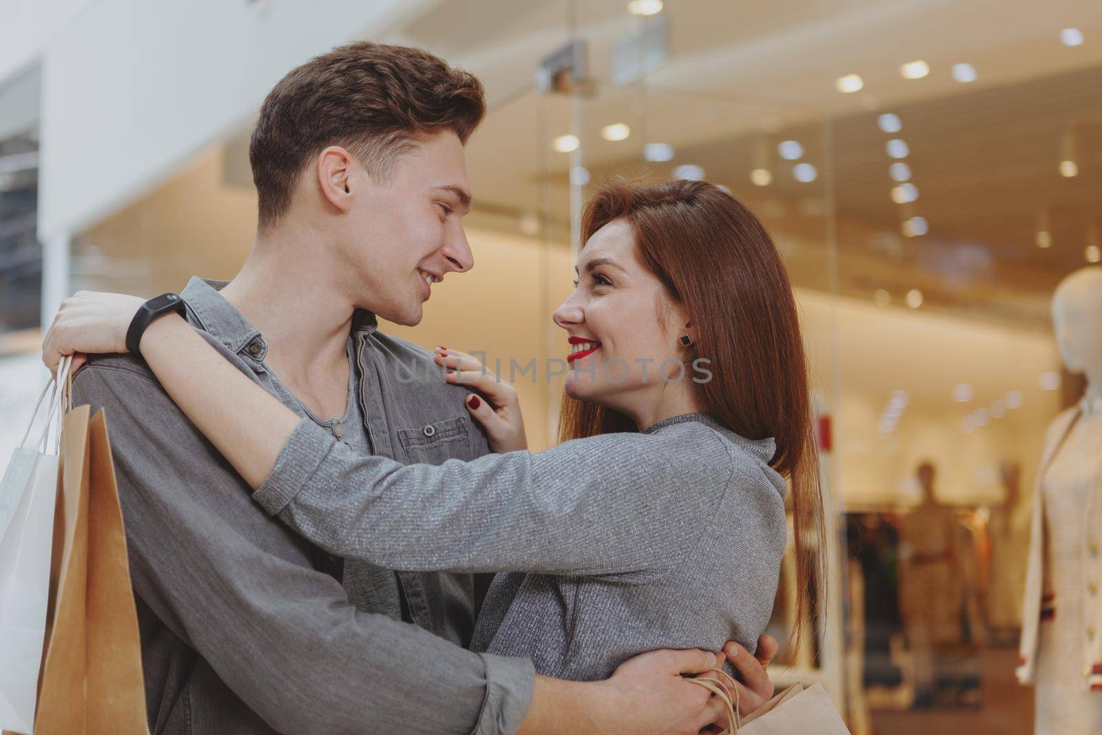 Happy young couple hugging, smiling at each other at the shopping mall. Young man embracing his girlfriend while shopping at the mall together. Love, anniversary, couples concept