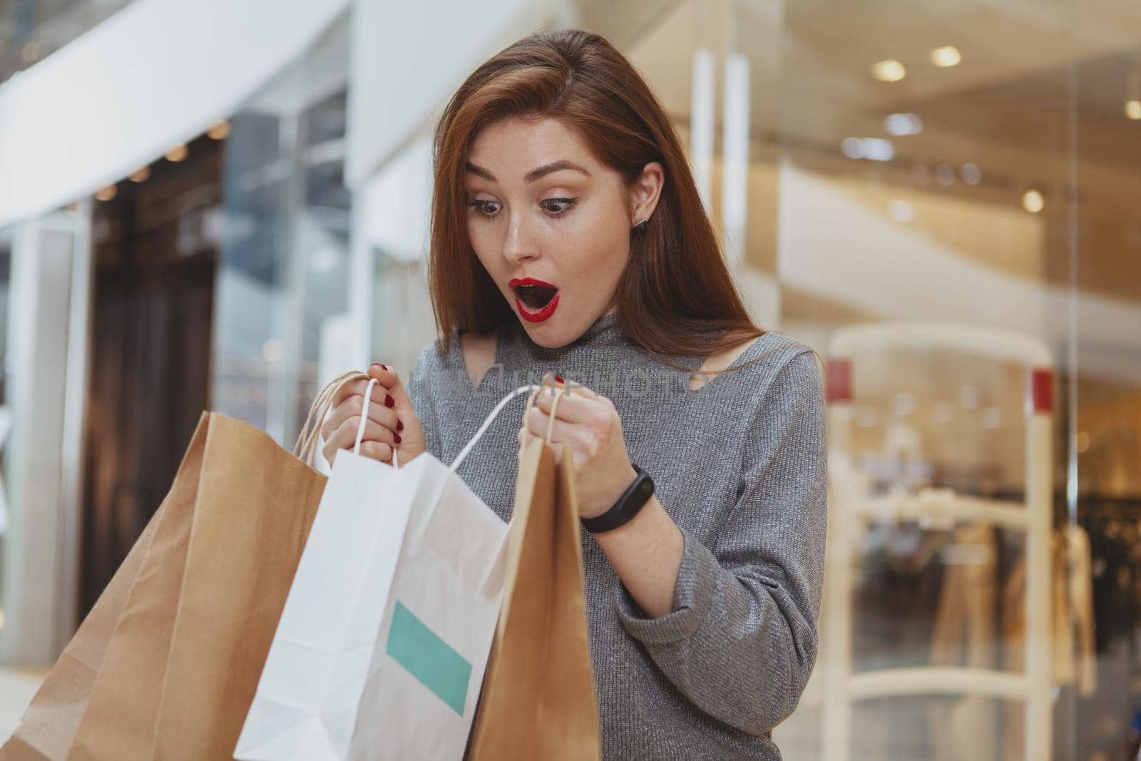 Beautiful overwhelmed woman looking inside a shopping bag at the mall. Attractive female cusotmer looking surprised, checking out her purchase in a paper bag, copy space. Sale, low price concept
