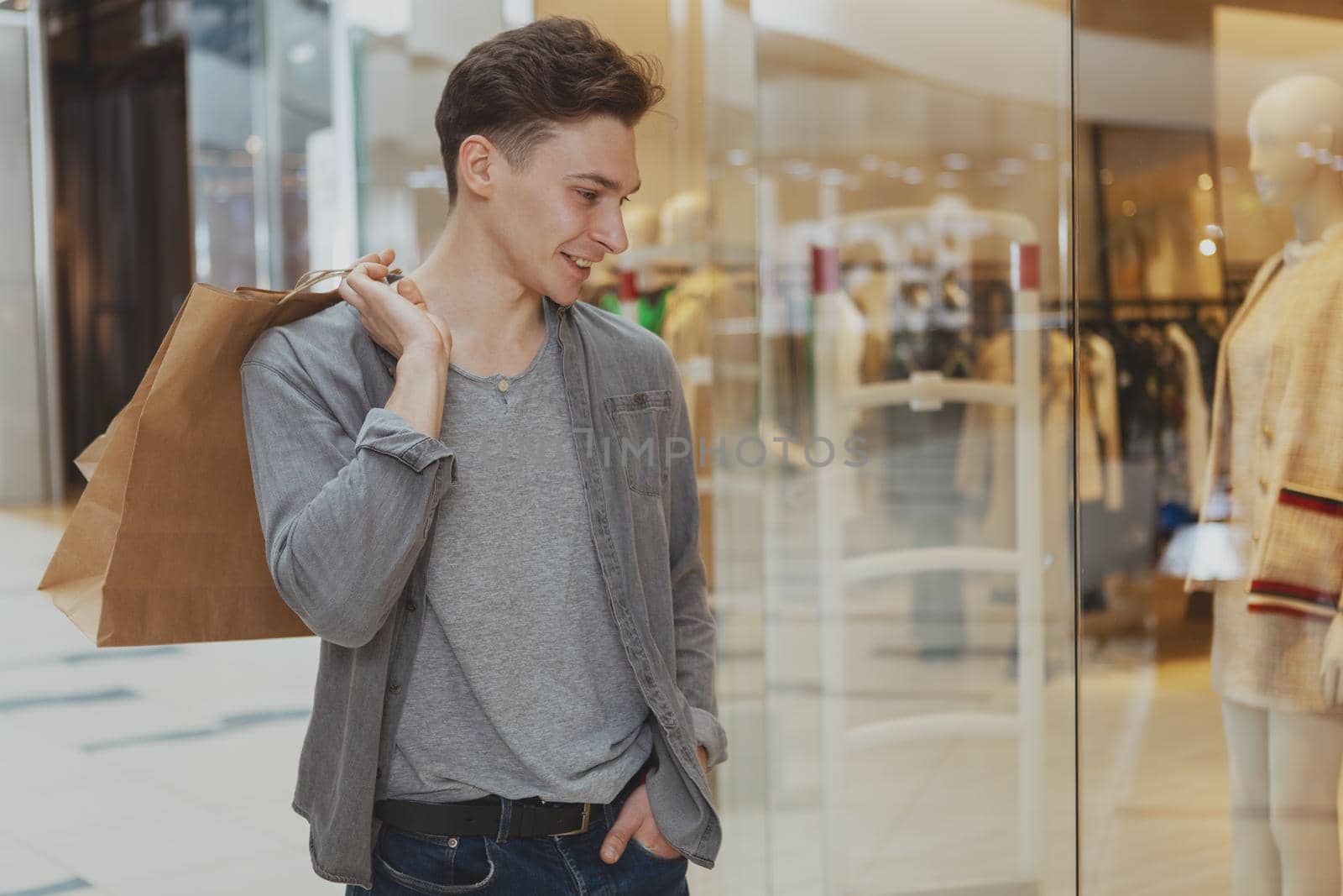 Attractive young man smiling, looking at the store showcase, walking at the shopping mall. Handsome male customer enjoying shopping at the mall, copy space. Lifestyle, leisure concept