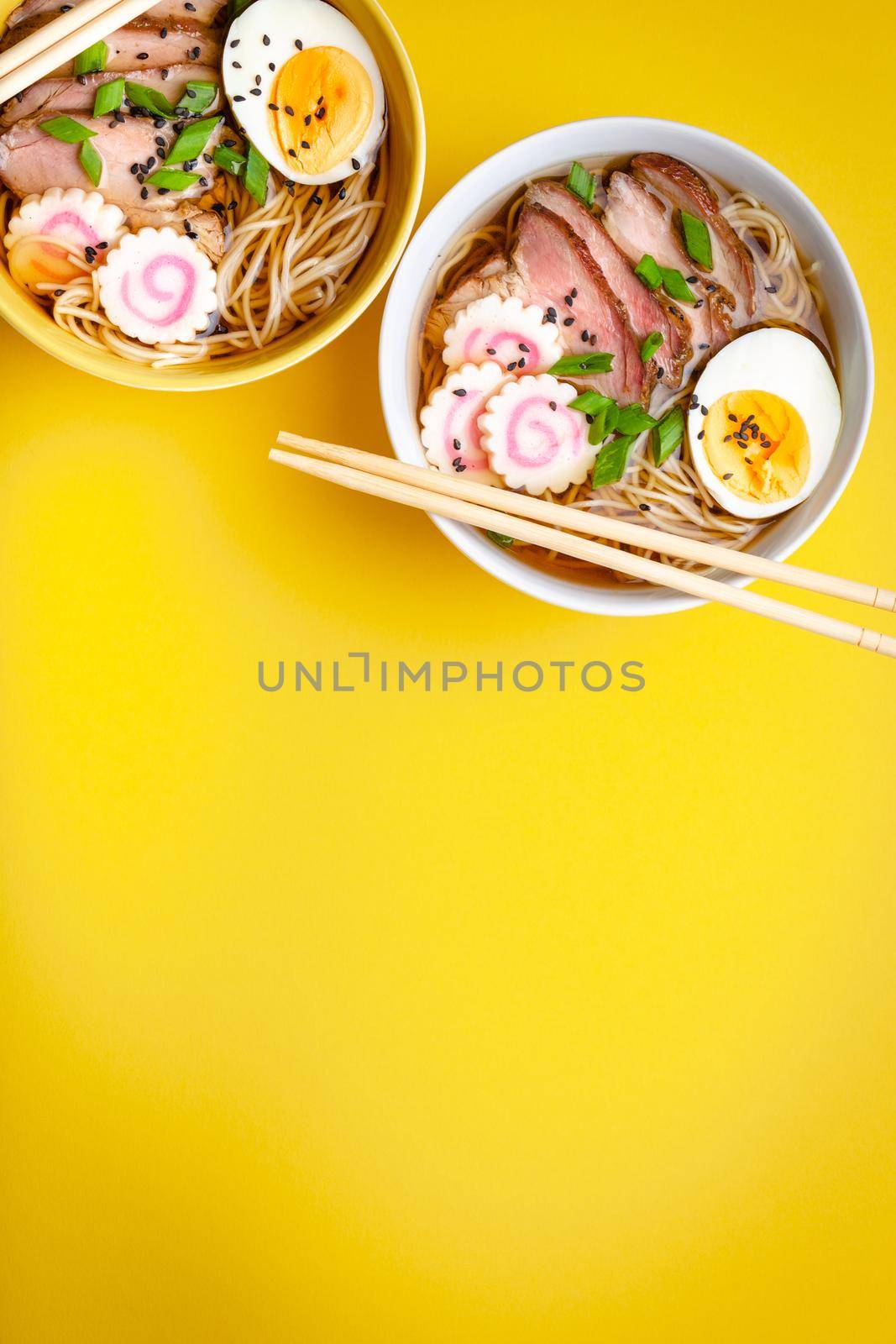 Two bowls of Japanese noodle soup ramen with meat broth, sliced pork, narutomaki, egg with yolk on pastel yellow background. Traditional dish of Japan, top view, close-up, space for text