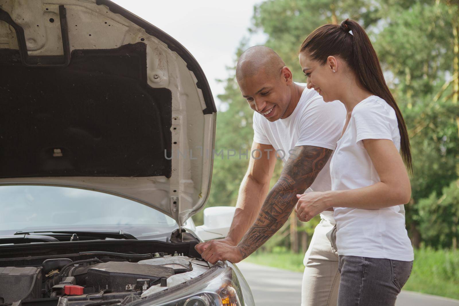 Young mixed couple laughing, looking under the hood of their broken car on the side of the road, copy space. Handsome African man examining engine of his broken car on a roadtrip with girlfriend