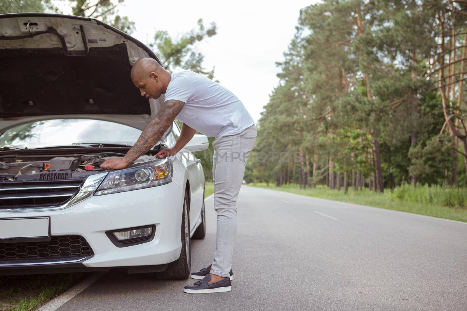 Full length shot of a young African man examining engine of his broken car on the side of the road, copy space. Male driver looking under the hood of his broken aute during roadtrip