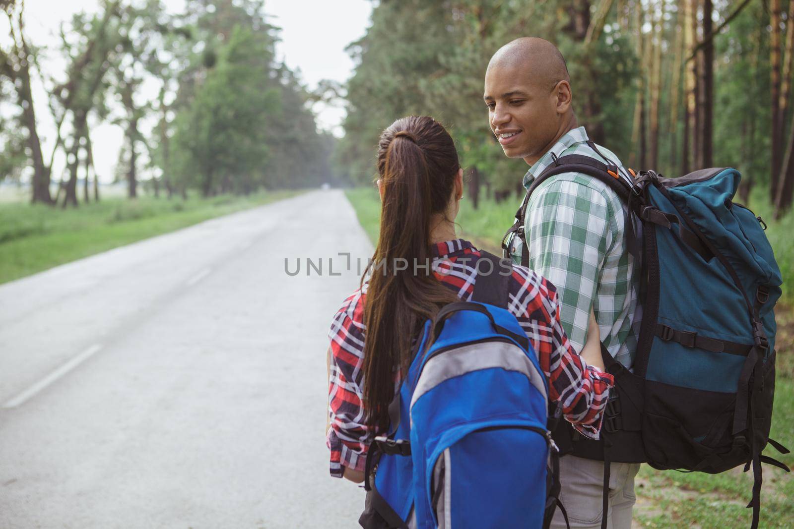 Handsome African man with backpack smiling at his girlfriend while hitchhiking on the countryside road, copy space. Young couple travelling together