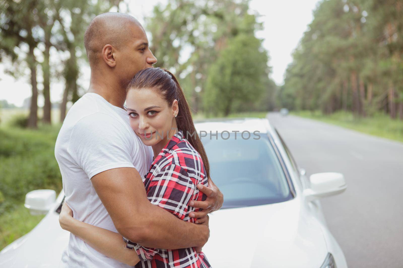 Beautiful young woman smiling to the camera, hugging with her boyfriend near the car on the road, while travelling together. Lovely multiracial couple on a roadtrip, cuddling near their automobile