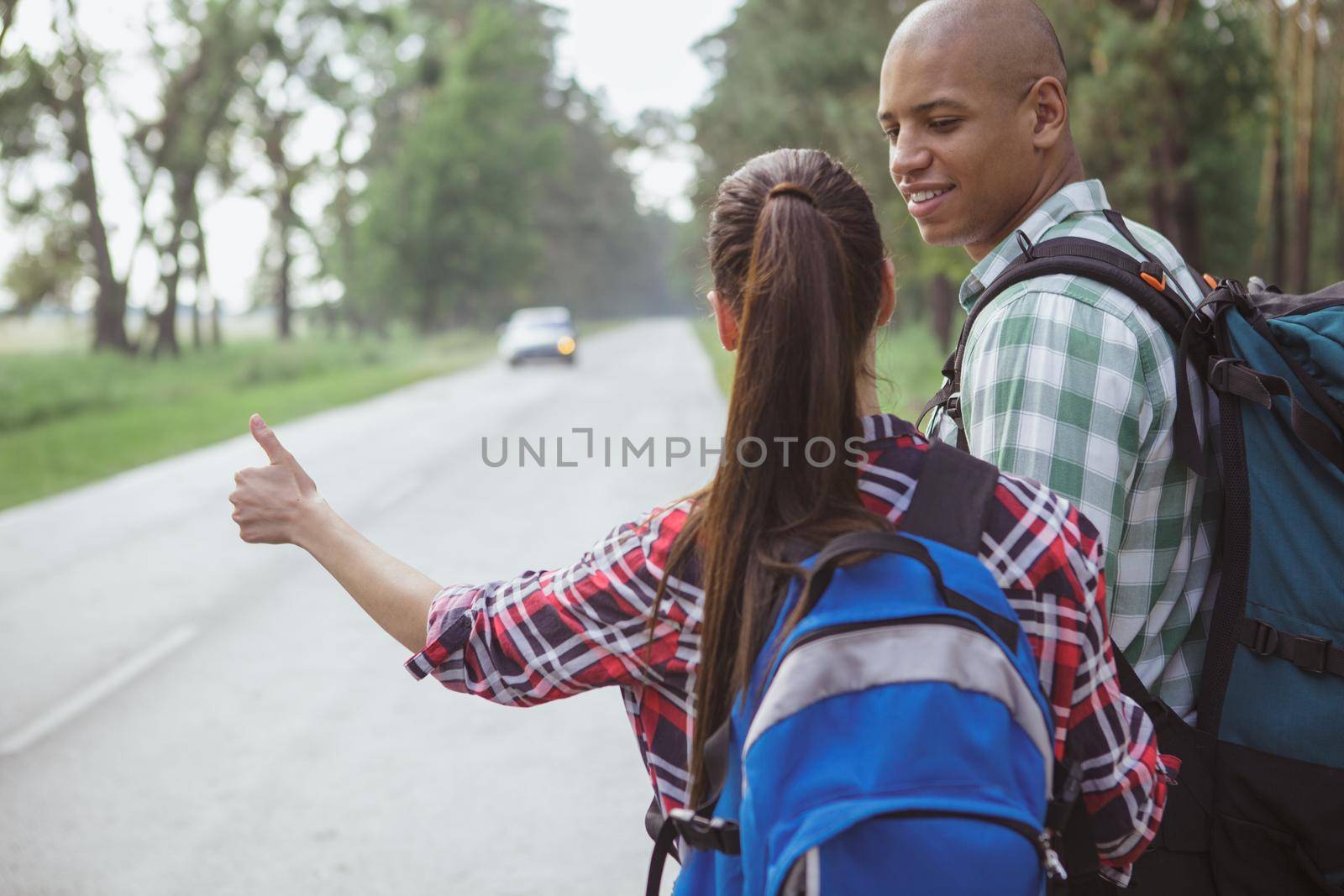 Rear view shot of a couple hitchiking on the countryside road in the forest, copy space. Cheerful handsome African man smiling at his girlfriend while backpacking together