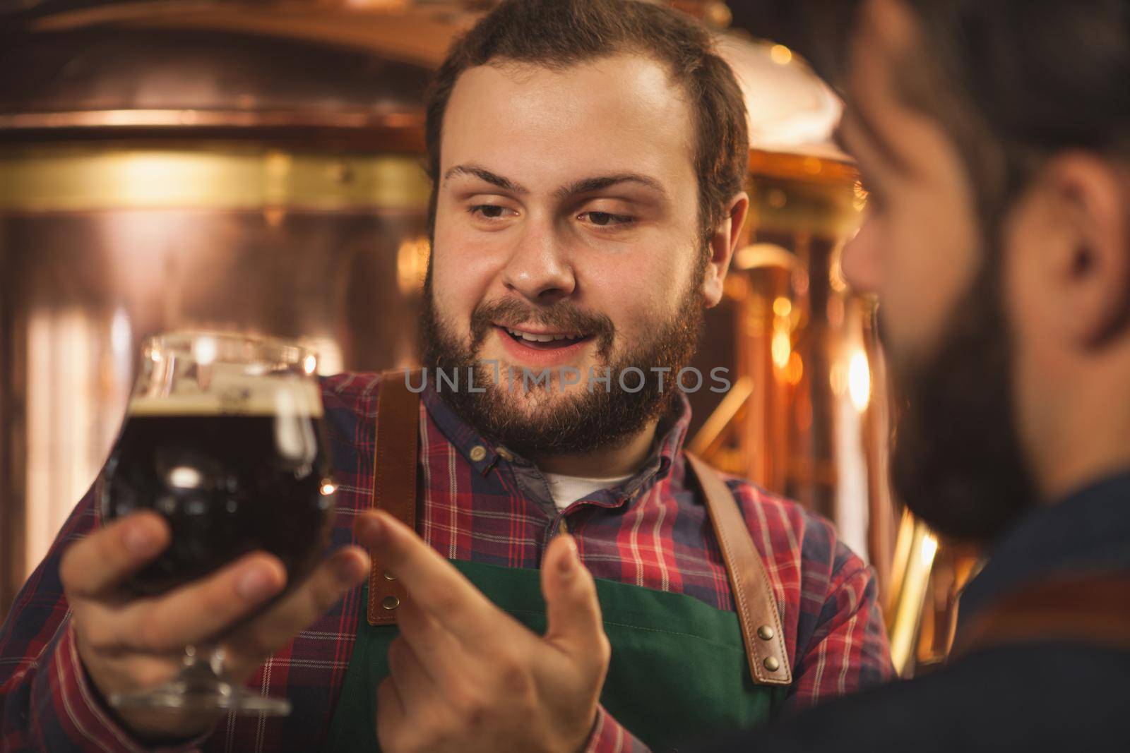 Close up shot of a cheerful bearded brewer smiling, talking to his colleague, examining dark beer in a glass. Two brewers producing craft beer at their microbrewery. Manufacturing, small business concept