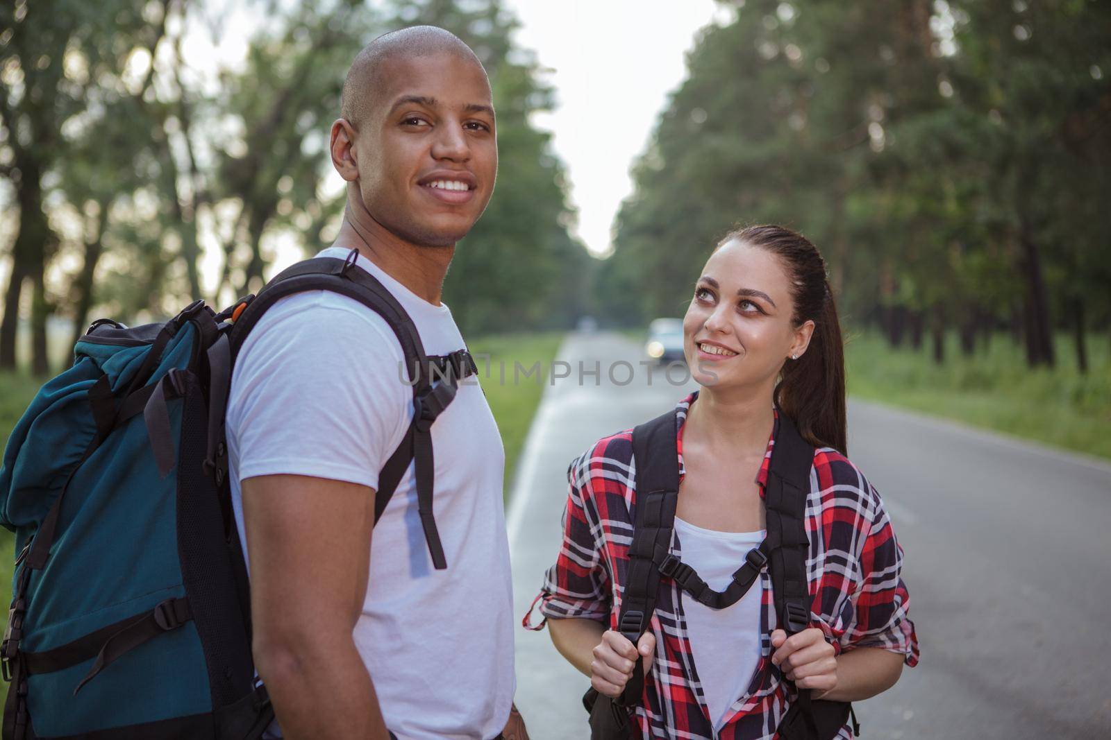Handsome African man and his charming girlfriend backpacking together, waiting for a car on the side of the road. Mixed couple hitchhiking on countryside road