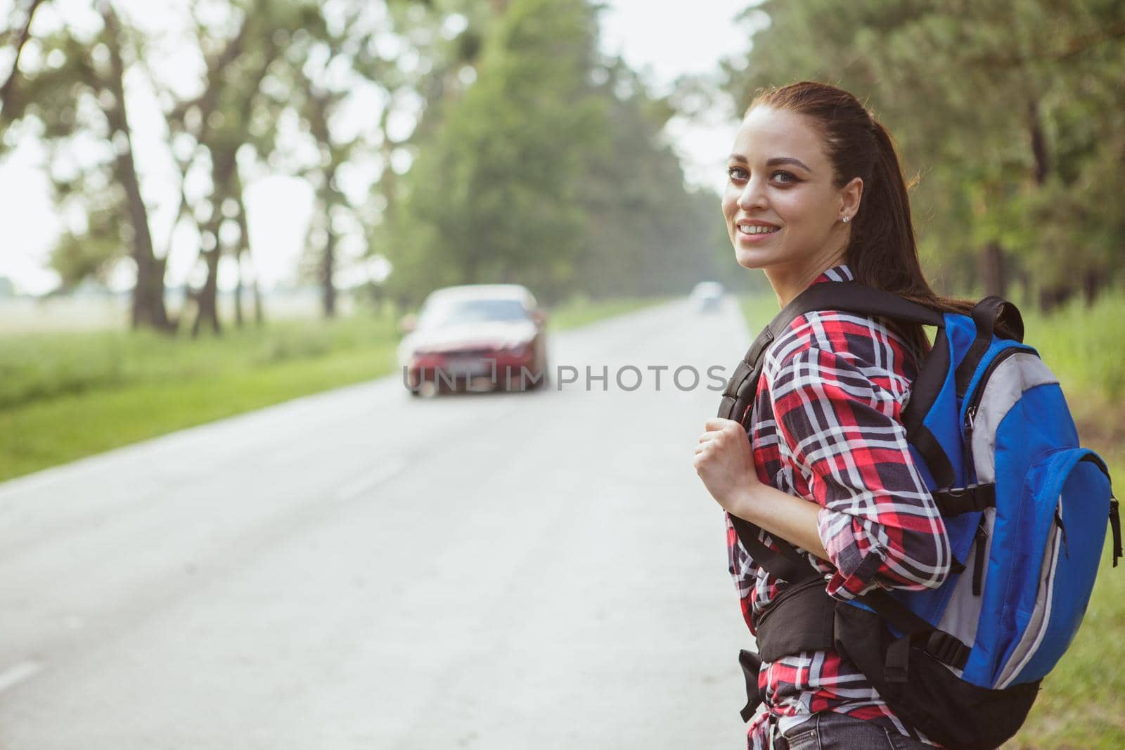 Beautiful happy young woman backpacking, smiling over her shoulder while hitchhiking on countryside road, copy space. Lovely female traveller with a backpack catching a car on the road in the woods