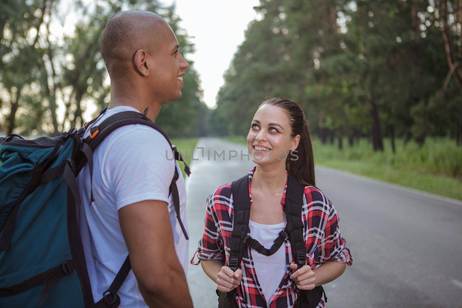 Lovely young woman with backpack smiling at her boyfriend, resting on the side of the road while backpacking and hitchhiking. Two hitchhikers on the side of the road.