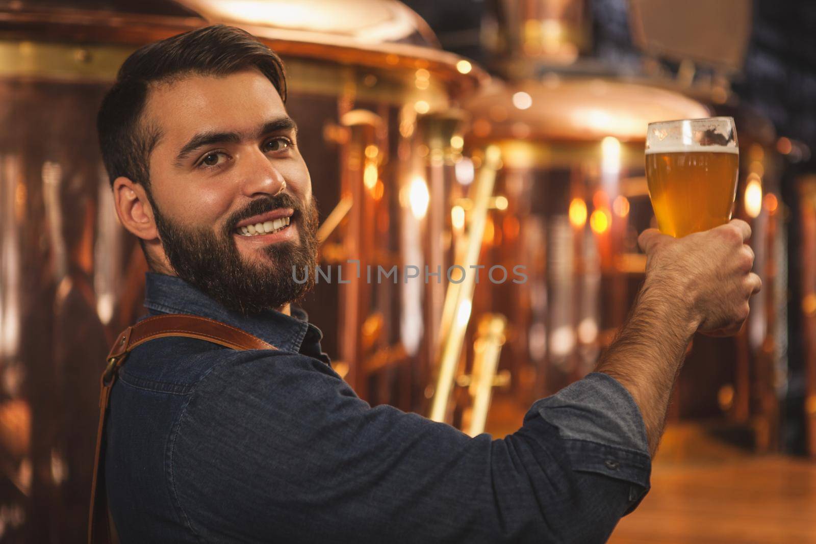 Happy bearded professional brewer smiling to the camera over his shoulder, holding glass of delicious beer, copy space. Attractive male bartender posing with a glass full of tasty beer. Bar, pub concept