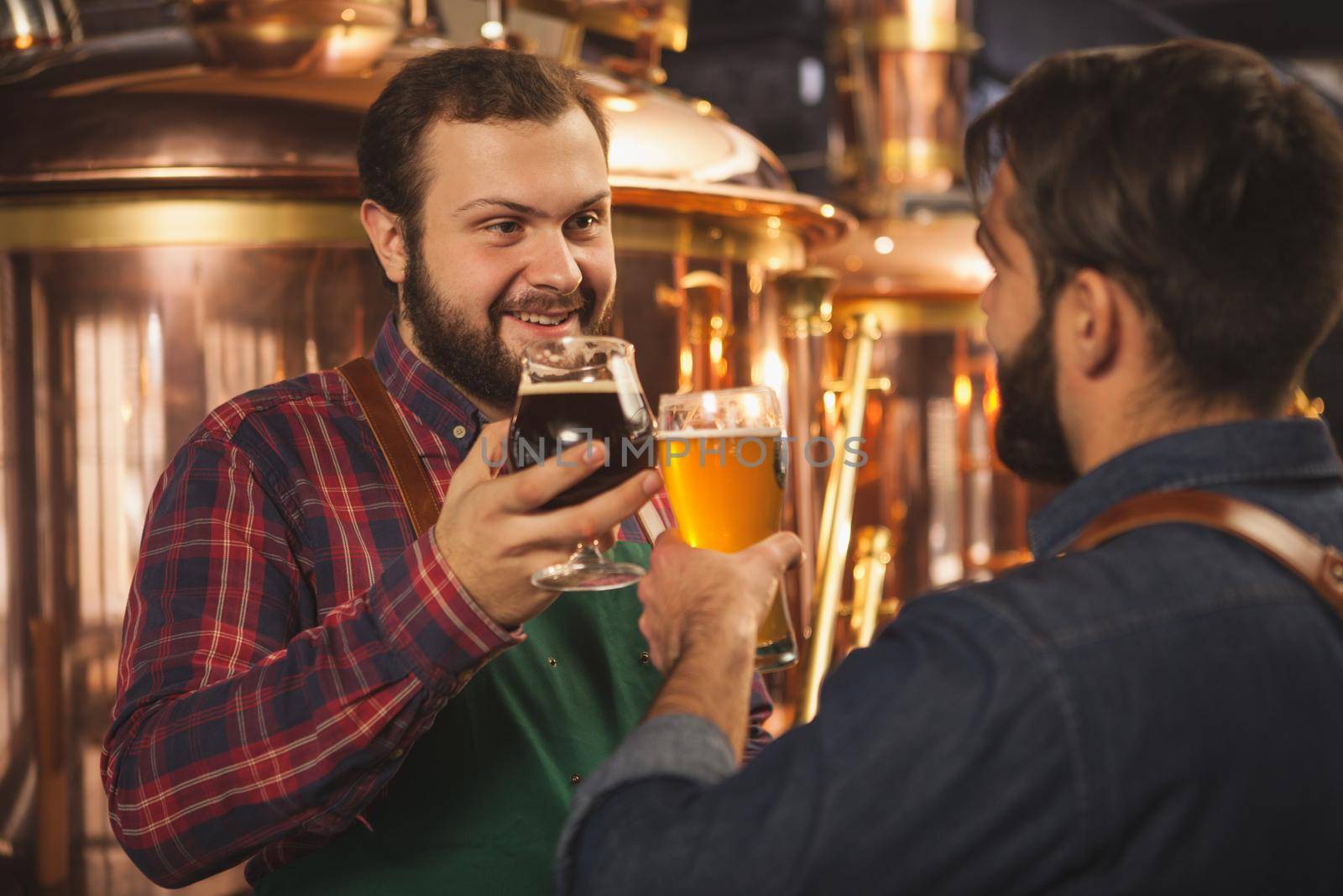 Bearded cheerful brewer talking to his assistant, working at the beer production production factory. Professional brewers clinking beer glasses, celebrating success. Small business, entrepreneurship concept