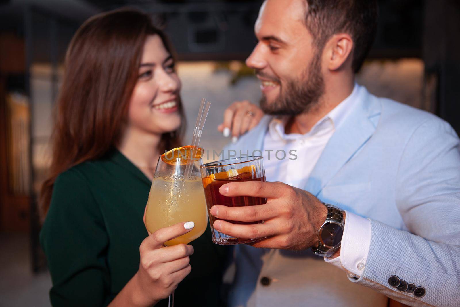 Cropped closeup of a happy loving couple clinking cocktail glasses, celebrating valentines day at the bar. Selective focus on drinks in hands of young couple