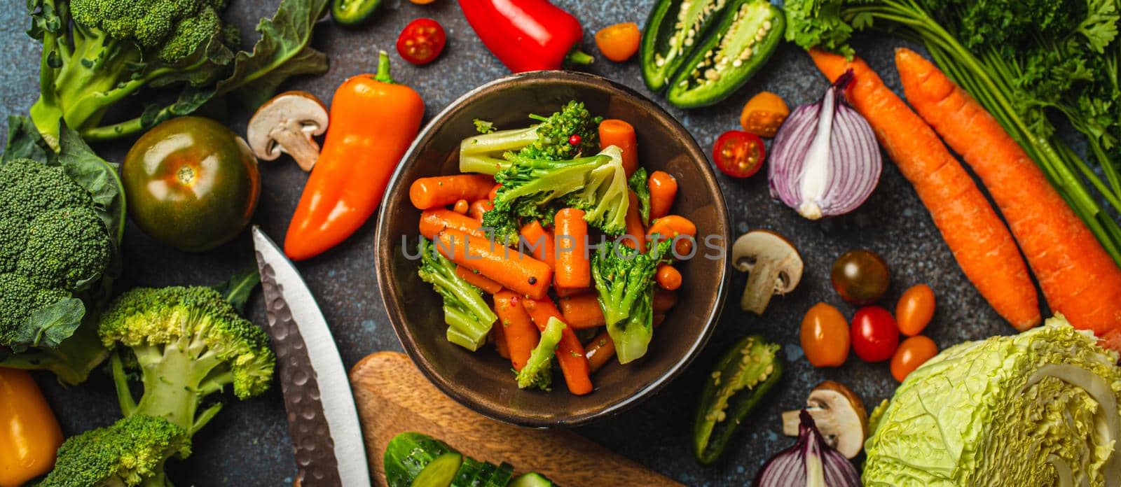 Steamed carrot broccoli salad in bowl and assorted cut fresh vegetables on rustic concrete table, wooden cutting board, kitchen knife from above, vegetarian or diet clean healthy food concept