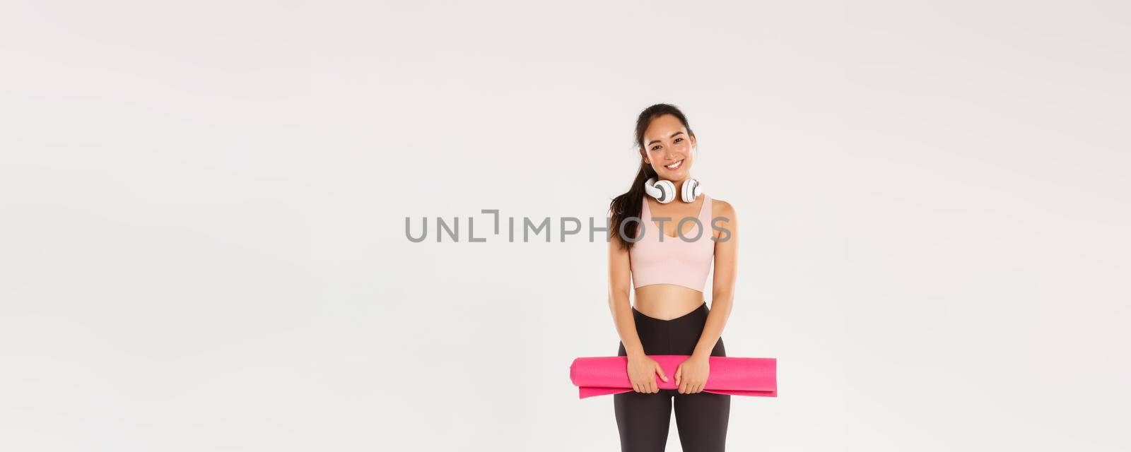 Full length of cute asian girl like fitness, holding rubber mat for exercises or yoga classes, standing in sportswear looking happy after productive training in gym, good workout, white background by Benzoix