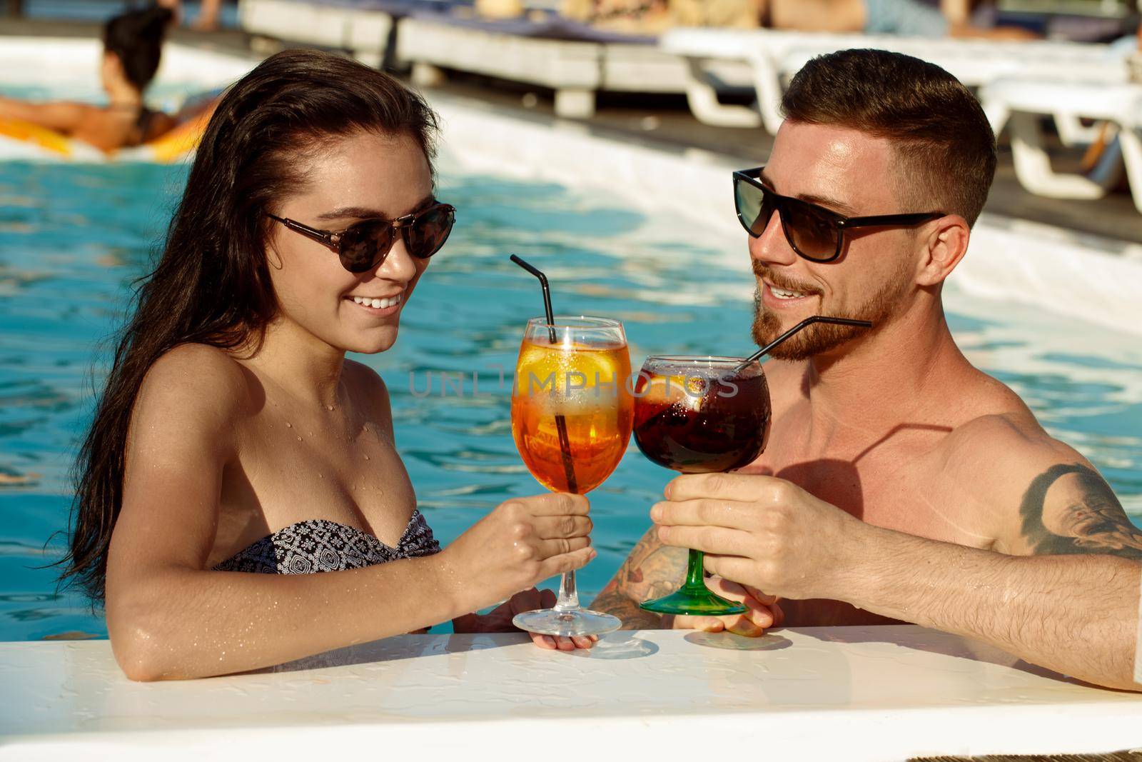 Happy loving young couple having drinks at the swimming pool. Attractive young woman and her handsome boyfriend smiling drinking cocktails at the pool. Tourism, travelling, summertime concept