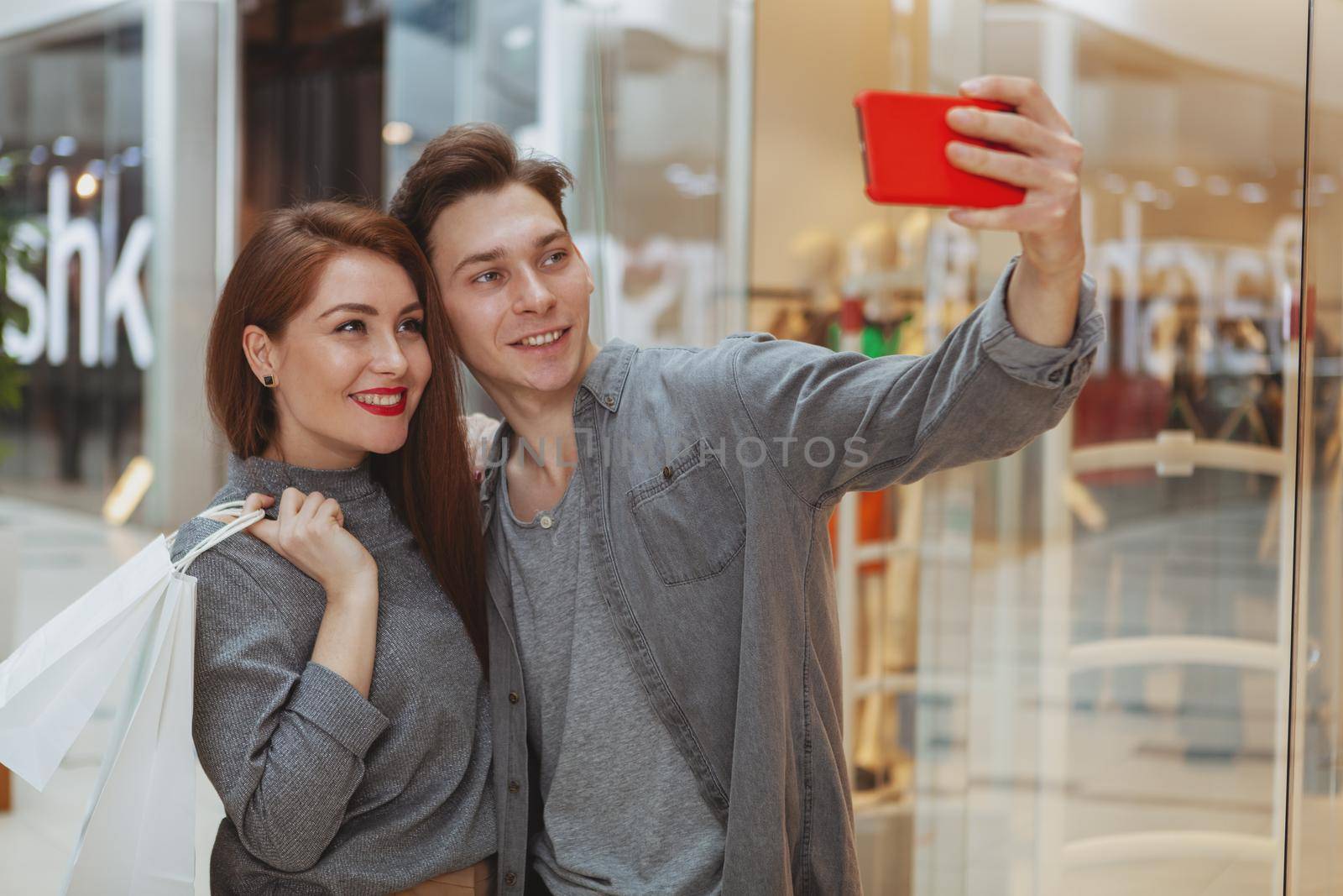 Adorable young loving couple using smart phone at the shopping mall, taking selfies together, copy space. Sharing, news, blogger concept