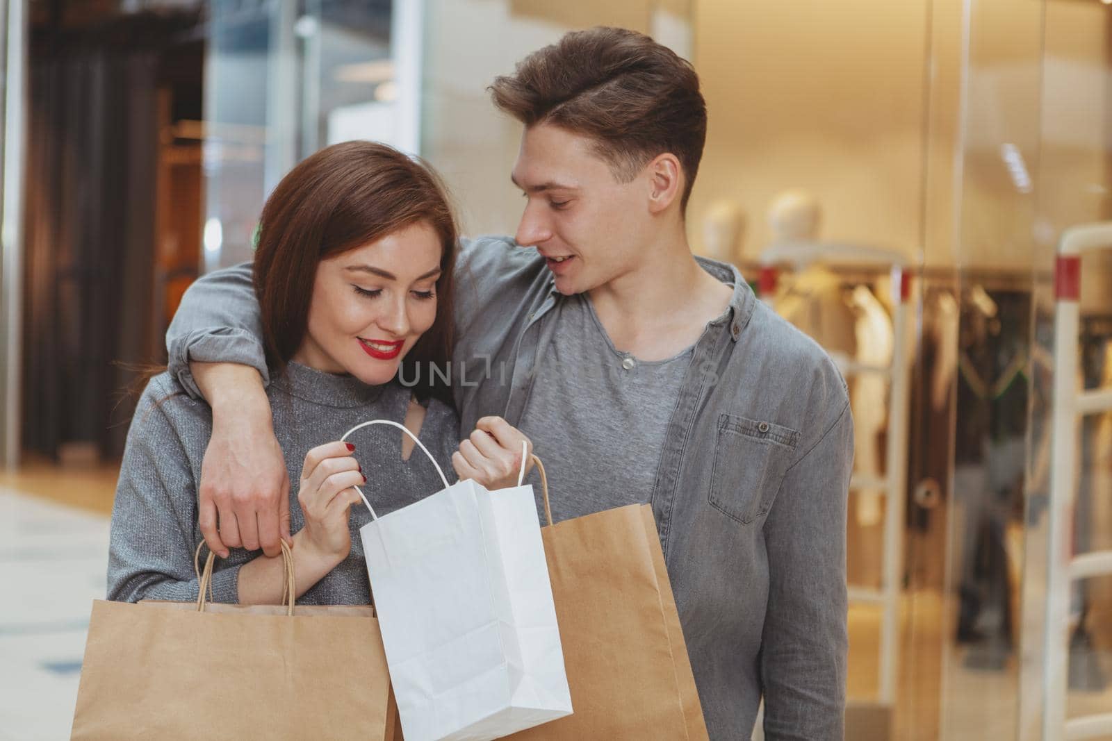 Handsome man smiling at his beautiful girlfriend while she is looking into a shopping bag. Adorable couple shopping together at the mall. Buying presents, celebrating anniversary concept